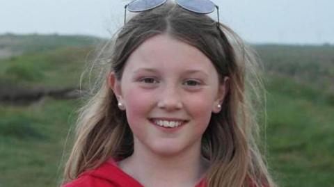 A close-up of Jessica Lawson, who is smiling for the camera. She has long, light-brown hair and wears a red top and has sunglasses on her head. She stands against a backdrop of green hills.