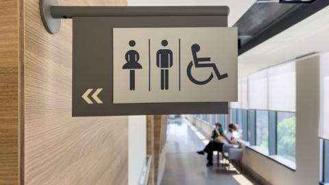 Toilet sign with corridor and people sitting on a bench in the background, with big windows.