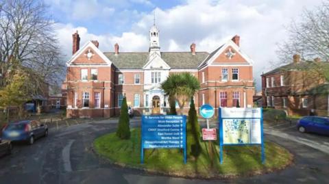 The front view of a red-brick building with a clock tower, surrounded by trees and parked cars. A blue sign in the foreground lists various services offered, and the building appears to belong to the North East London NHS Trust.