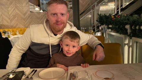 Michael Woods, 36, who has ginger hair and a short ginger beard, sits at a table in a restaurant with his arm around his son, six, who has short blonde hair. 