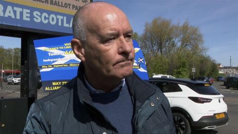 Tommy Sheridan, who has short grey hair, in front of a branded billboard next to a road