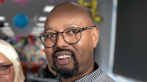 Abdullahi Abdi wearing a blue and white checked collared shirt underneath a waffle patterned grey jumper. He is wearing black framed glasses, has a shaved head and a dark goatee style beard, and is smiling at the camera.