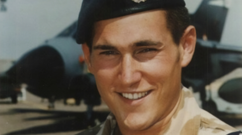 A close up picture of a smiling man with a beret. It's a head-and-shoulders image but is cropped tightly so you can see his facial features. He is smiling and standing in front of a military plane which appears in the background.