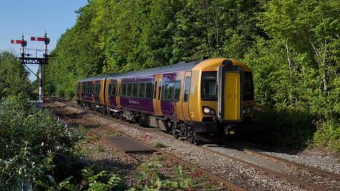 A purple and yellow West Midlands Railway train travels along a track through a heavily wooded area, with trees and plants either side of the track. Two red and white signal arms are visible on the left of the track.