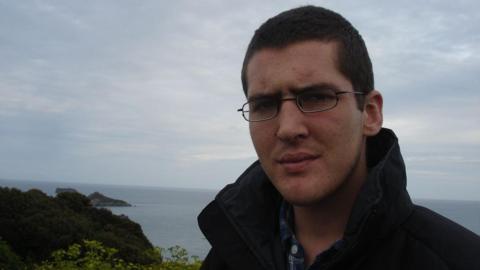 A man wearing rectangle black glasses and a high neck black coat with the sea and cliffpaths in the background.