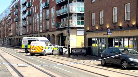 Blue and white police tape stretches across a city centre street. behind the tape, two police vans and a police car are parked , blocking the junction to a side street. The bar is a three-storey red brick building with floor to ceiling windows at ground level and two rows of four standard windows on the floors above. The next building along the street is a modern seven-storey block of apartments with glass-fronted balconies. It is a sunny day with a blue sky.
