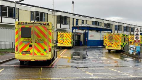 The front of the Queen Elizabeth hospital in King's Lynn showing the ageing cladded two storey building, built with reinfoced autoclaved aerated concrete and multiple emergency ambulances queuing outside 