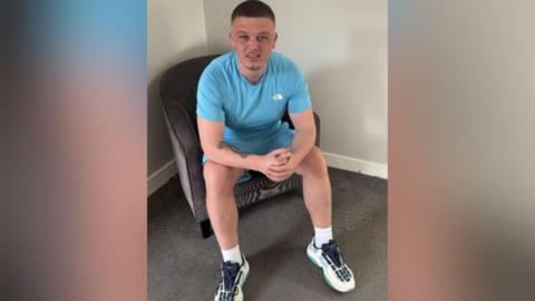 A man with short hair sitting on a chair wearing a blue top