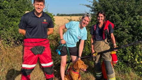 A man and two women standing beside a brown dog on a grassy path next to a field, with trees and hedges in the background. Two people are wearing firefighters uniform and a woman is holding the dog’s lead.