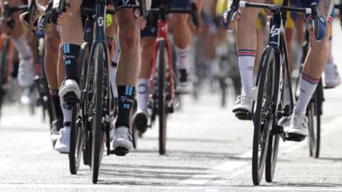 A large group of cyclists are riding along the road. They can only be seen from the legs down. 