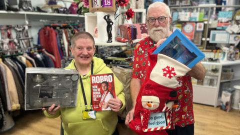 A man and a woman are standing in a charity shop. The woman is wearing a green hoodie, and is holding a gift set of jars and a book. The man is wearing a red Christmas t-shirt, and is holding a stocking with a fragrance gift set peeking out of the top.