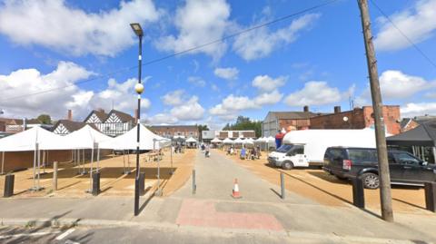 Rows of white canopies on a gravel area with a path down the centre, seen on a sunny day. White clouds float in the sky. A white van and black estate vehicle are parked on the right, and people can be seen in the distance.