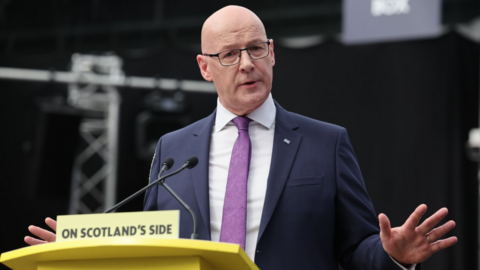 John Swinney, who is bald with glasses, at a podium