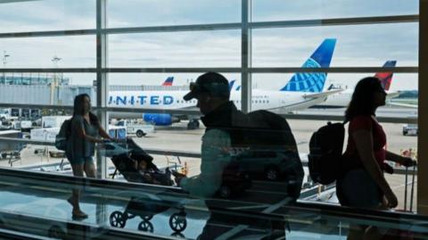 People at an airport walking past glass windows with planes in the background.