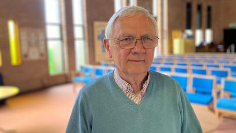 Philip wearing blue v-neck jumper and a checked shirt inside - standing inside the main prayer hall of the church- with blue chairs in the background