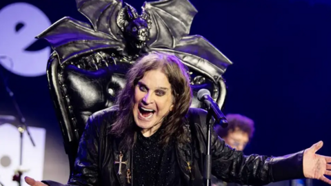 Ozzy Osbourne dressed in a black leather jacket sat on a black throne with a microphone in front of him. He has his mouth open wide as he shouts at the crowd, gesturing with his left hand. A microphone is in front of him, and a cross is hanging off his jacket.