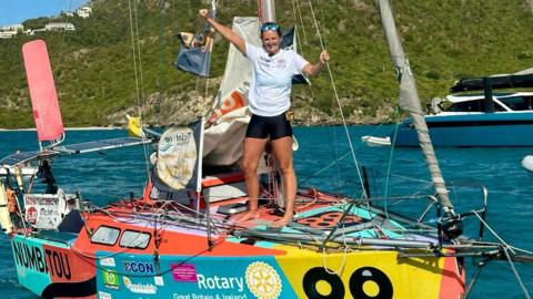 A woman standing on the deck of a small, brightly decorated sailing vessel. She is standing barefoot near the bow, with both arms raised in a celebratory gesture. She is wearing a white short‑sleeved shirt, dark shorts, and reflective sunglasses. The boat is covered in colourful graphics and sponsor logos. The hull features bold, vibrant patches of red, yellow, turquoise, and orange.
Visible text includes: “NUMBATOU” on the bow. Rotary Great Britain & Ireland along the side, accompanied by the Rotary emblem. The sail has multiple stickers or patches and some printed text, though parts are crumpled or folded, making them partially unreadable. A prominent pink rudder or trim tab is visible at the stern. The boat is floating on calm, clear turquoise water. The backdrop features lush green hills with several white buildings scattered across them. To the right, a larger motor yacht is moored, adding to the coastal marina atmosphere.