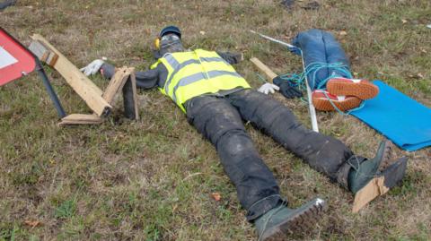 A broken scarecrow wearing a yellow hi-vis jacket lies on a grassy verge, it is surrounded by debris and the legs of another scarecrow.