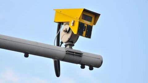 A yellow average speed camera on top of a grey pole against a blue sky