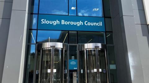 The picture shows the entrance to Slough Borough Council's offices. White letters spell out "Slough Borough Council" against blue tinted glass. There are two revolving doors.  