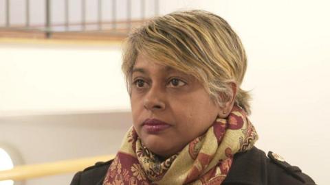 A woman, pictured from the shoulders up, wearing a gold and red scarf and a black jacket. She has brown and blonde hair. 