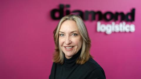 A woman with blonde hair. She is wearing a black jumper. The background behind her is pink. 