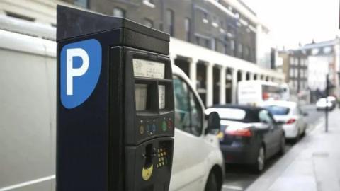 The side of a black parking metre. There is a blue sticker with a white P on it. In the background is a white van parked behind a number of cars in a street