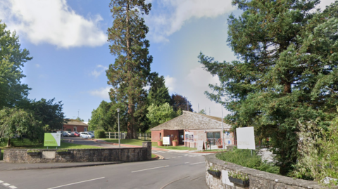 A Google Street View screenshot of the entrance to HMP Leyhill showing a one storey building behind a low stone wall surrounded by trees.