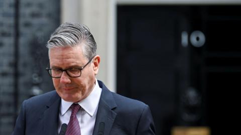 A man, Keir Starmer outside number ten Downing Street. He is wearing a dark suit jacket, a white shirt and a patterned burgundy tie. His head is tilted forward in the direction of the stand and microphone from which he is making his speech