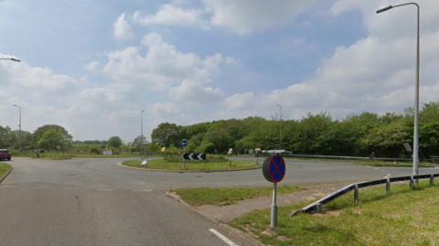 A roundabout at junction 16 of the A12 between Galleywood and Stock. There is grass on the middle of the roundabout as well as the verges either side, and there are grey barriers and green trees in the background.