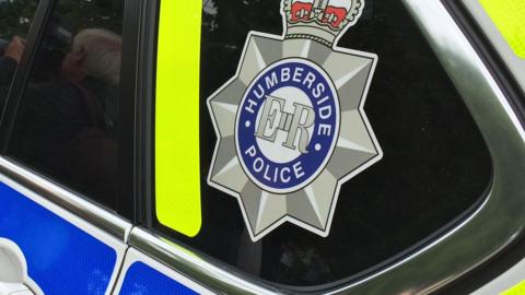 A close up of the back of a Humberside police car showing the logo with ER at the center.  The emblem is a star badge, topped with a crown, and a blue circle in the centre saying Humberside police.