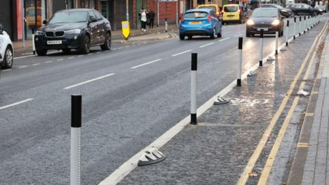 A section of the Linthorpe Road cycle lane which is marked by a solid white line and black-and-white poles. Cars are driven on the road to the left of the cycle lane.