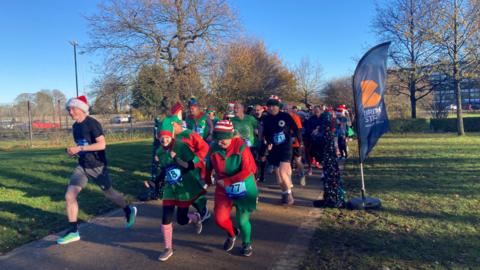 Runners dressed in elf outfits are running through the park.  The sky is bright blue and the sun high lights the colours well.