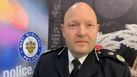 Mr Guildford is wearing his uniform and is stood by a West Midlands Police sign. He has a shaved head.