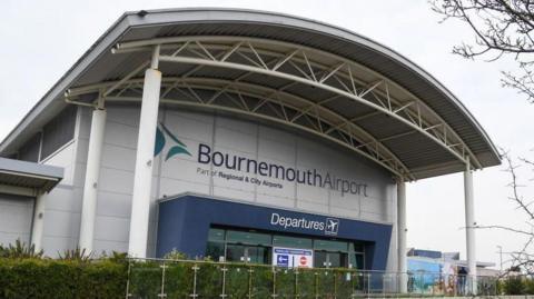 Bournemouth Airport Departure building - a grey building with a large portico held up by a number of columns and a blue surrounding to the glass entrance and a green hedge at the front.