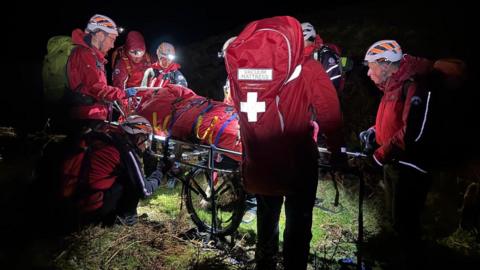 A group of mountain rescue team members huddle around a casualty who is being transported on stretcher. It is dark but the casualty is being lit up by head torches. The team wear emergency waterproof clothing which is red.