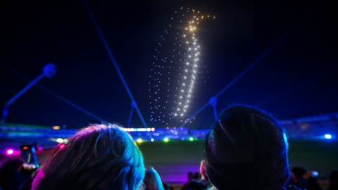 Drones make the shape of a penguin in the sky. Spectators are in the foreground looking up at the display.