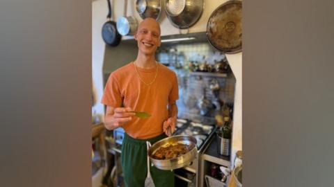 Elijah A.M in an orange tshirt and green joggers holding a pan of food towards the camera with something in his other hand that looks like a leaf