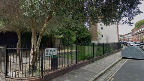 View of a small and basic fenced park with trees and paved paths. It is located on a residential street, a grey bike storage unit is visible outside the park.