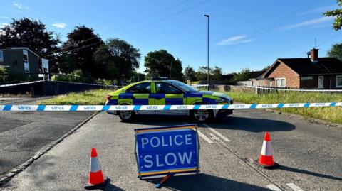 A police cordon in the middle of Thurncourt Road. A blue 'police slow' sign sits between two cones in front of a police car parked inside a taped-off cordon.