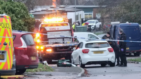 A police van, a maroon people carrier, a car removal truck and a white car parked in a estate. Several police officers are talking to each other and police tape has been set up.