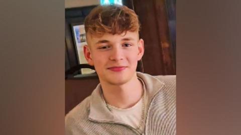 George, a teenager with curly blond hair shaved at the sides and long at the top, smiles into the camera while wearing a beige coloured fleece open at the neck.