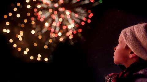 A child wearing a woolly hat stands watching fireworks going off.