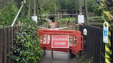 A red and white sign which says footpath closed stands in front of a red plastic barrier blocking entry to a footbridge leading away from the camera towards the stone entrance to a tunnel which can be seen in the background among trees.