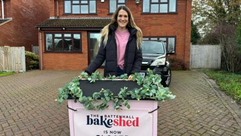A woman with long blonde hair is pictured wearing a pink jumper and a black coat whilst stood at the end of a driveway in front of a small pink cupboard that comes up to her waist. The roof of the cupboard has plastic ivy around it with a white sign that reads "The Battenhall Bake Shed". 