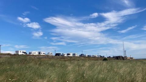 A row of campervans silhouetted against a blue sky in front of a field of long grass are parked along a narrow road 