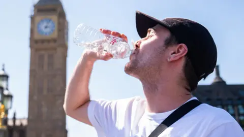 A man drinks from a water bottle with a sunny sky overhead and a blurred view of the Westminster skyline in the background