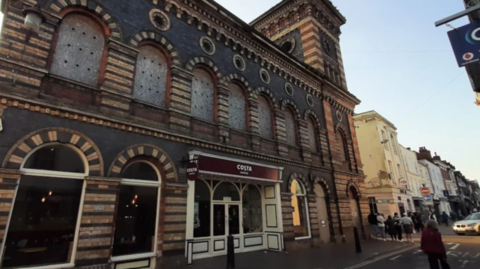 A large building with large windows and a decorative brick front and a small red sign with the word Costa on it