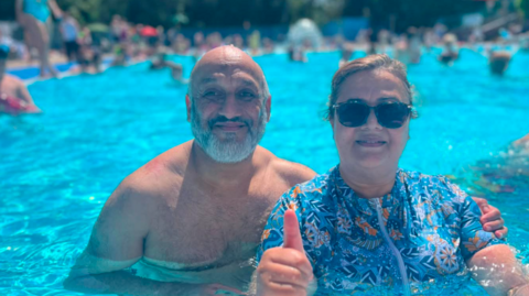 A man with a bald head and grey beard next to a woman with brown hair and a patterned blue top with her thumb in the air. Both are half submerged in a blue pool