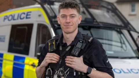 PC Thomas Fairbrother‑Wilcock has short dark brown hair and stands in full uniform in front a police van.